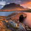 The image is well-composed with a clear focal point on the stone bridge. The lighting is excellent, capturing the warm tones of either sunrise or sunset. It is sharp and detailed, showcasing the texture of the stone and the surrounding landscape. The colors are accurate and vibrant, contributing to a photo-realistic feel. Key elements, such as the bridge and the distant mountains, are in focus. Overall, the image excels in composition, lighting, sharpness, color accuracy, and photo-realism.