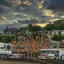 The image is well-composed, capturing a town nestled in a landscape with a cloudy sky. The lighting is good, highlighting the buildings and surrounding greenery. The sharpness is acceptable, allowing for decent detail. The colors appear accurate, and the image has a photo-realistic quality. The key elements, such as the buildings and the structure on the hill, are in focus. There are no unwanted elements present, such as watermarks or cluttered environments. The composition uses the rule of thirds effectively, placing the main subjects in visually appealing positions. The dynamic range is well-handled, with details visible in both the highlights and shadows. The image presents a balanced and aesthetically pleasing view of the town and its surroundings.