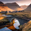 The image is well-composed, showcasing a scenic landscape with a stone bridge over a river, a village nestled in the valley, and mountains in the background. The lighting is warm and inviting, likely captured during sunrise or sunset. The image is sharp and the key elements, such as the bridge and the village, are in focus. The colors appear accurate and the photo-realism is high. The reflection in the water adds to the overall aesthetic appeal. Strengths: Well-composed, well-lit, sharp, color-accurate, photo-realistic, key elements in focus. Weaknesses: None apparent.