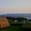 The image depicts a landscape with a cabin in the foreground, an ocean, and mountains in the background. The composition is decent, but the cabin is slightly dark. The lighting is soft, likely taken during dusk or dawn. The sharpness is acceptable, and the colors appear accurate. It has a photo-realistic quality. The key elements, such as the cabin and the distant mountains, are reasonably in focus. The image is well-composed, and the overall scene is appealing. The image could be improved with better lighting on the cabin.