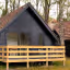 An exterior shot of modern black cabins with traditional tiled roofs, nestled among trees. A wooden deck and railing are visible in the foreground, with outdoor furniture on the deck.