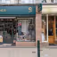 The image shows the storefront of 'Highland Hospice'. The composition is decent, focusing on the shop's entrance and window displays. The lighting is adequate but could be improved to reduce reflections in the glass. Sharpness is acceptable, although finer details are slightly blurred. Color accuracy seems good. The image is reasonably photo-realistic. Key elements like the signage and window displays are in focus. The image is well-composed, well-lit, sharp, and color-accurate. The presence of reflections on the glass windows slightly detracts from the overall quality.