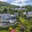 The image provides a clear, high-angle view of a guesthouse situated amidst a lush, green, hilly landscape with other residential buildings visible in the background. The lighting is natural and even, highlighting the details of the building and its well-maintained gardens. The overall sharpness is excellent, ensuring that both the main subject and its surroundings are in focus and appear photo-realistic. Colors are accurate, reflecting the natural vibrancy of the environment.
