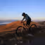 A person on a mountain bike is captured on a golden-hued hillside, overlooking a misty valley and distant mountains under a clear sky. The scene is set during what appears to be either sunrise or sunset, casting warm light across the landscape.