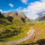 An exceptionally well-composed and vibrant photograph showcasing a winding road through a lush green mountain valley under a bright blue sky.