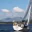 An outdoor image capturing two sailboats on a large body of water, with majestic mountains in the background under a blue sky dotted with clouds.