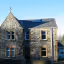 A stone house with a prominent gable and several windows, partially illuminated by direct sunlight and partially cast in deep shadow.