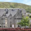 An outdoor photograph of a large, traditional stone hotel building with multiple dormer windows and gabled roofs, set against a green, hilly backdrop under a bright blue sky. A hedge and parked cars are visible in the foreground.