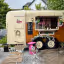 The image depicts a food truck called 'Bloom and Graze'. The composition is acceptable, but the presence of a pink trash can is visually distracting. The lighting is adequate, but the sharpness could be improved. The colors appear accurate. The image is reasonably photo-realistic. The key elements, such as the food truck itself and the menu board, are generally in focus. However, the overall impression is somewhat diminished by the trash can and the slightly cluttered environment.