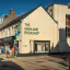 An exterior view of 'The Highland Bookshop' on a sunny day, with people walking on the cobblestone street in front of it. The building is a prominent feature, rendered in light yellow with green trim and text.